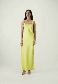 Vestido de cetim amarelo até ao chão, com alças finas e decote quadrado. Textura suave, design ajustado, combinado com sapatos de salto bege.
