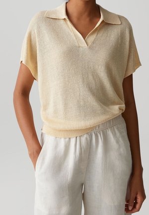 Femme portant un polo en maille beige clair à manches courtes, rentré dans un pantalon large en lin blanc taille haute, une main dans la poche.
