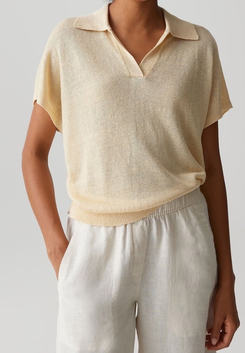 Femme portant un polo en maille beige clair à manches courtes, rentré dans un pantalon large en lin blanc taille haute, une main dans la poche.