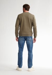 Sudadera verde oliva con puños y dobladillo acanalados, combinada con unos vaqueros azules de lavado desgastado y bolsillos traseros detallados, usados con zapatillas blancas.