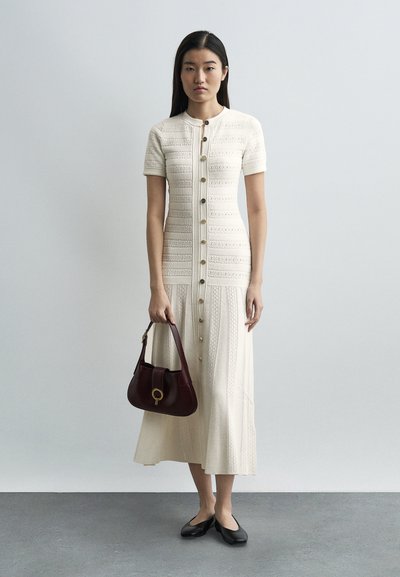 Γυναίκα με κρεμ πλεκτό φόρεμα με κουμπιά, κρατώντας σκούρα καφέ τσάντα χειρός, φοράει μαύρα φλατ παπούτσια, στέκεται μπροστά σε απλό γκρι φόντο.