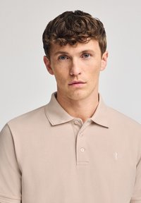 Camisa polo beige hecha de tela texturizada, con una tapeta de tres botones y un pequeño logo bordado en el lado izquierdo del pecho.