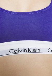Fialová športová podprsenka z bavlny s širokým bielym elastickým pásom s čiernym značkovým nápisom "Calvin Klein" na spodnej strane. Hladká textúra, zaoblený výstrih.