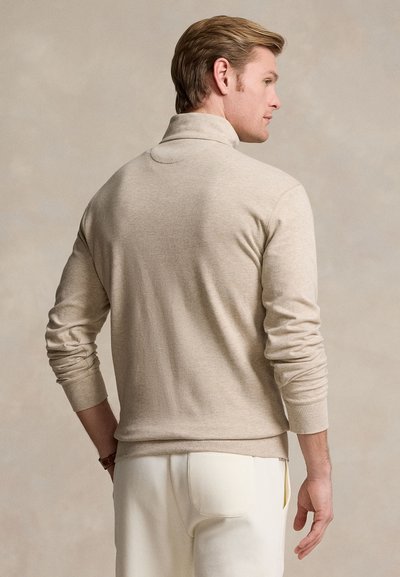 Hombre con cabello castaño claro visto de espaldas, vestido con un suéter de cuello alto beige de manga larga y pantalones blancos, sobre un fondo neutro.