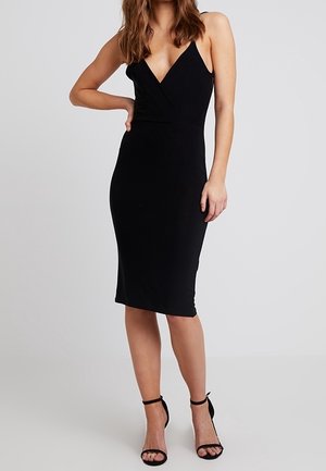 Femme portant une robe noire sans manches, longueur genoux, avec un col en V et des escarpins noirs à bride à la cheville, debout avec une main sur la hanche.