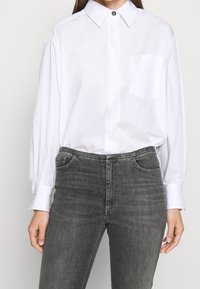 White long-sleeve shirt with a button-down collar, featuring a single chest pocket. Paired with dark grey fitted jeans, showing a smooth texture.