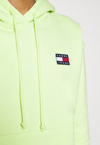Felpa verde neon con coulisse, caratterizzata da una toppa rettangolare ricamata nei colori blu navy, rosso e bianco. Tessuto morbido e tasca a marsupio.