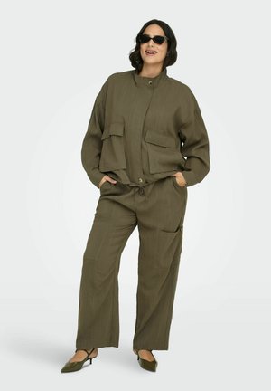 Kvinna iklädd oversized olivgrön jacka och byxor med fickor, svarta kattsyn-solitärglasögon och spetsiga kitten heel-skor, leende med händerna i fickorna.