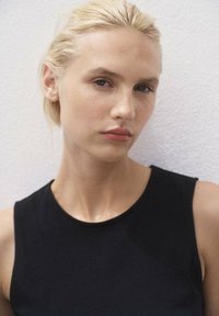 Model met kort blond haar en een lichte huid, gekleed in een mouwloze zwarte top. Neutrale achtergrond benadrukt haar gelaatstrekken.