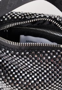 Zwarte gevlochten tas met metalen vierkante accenten en zilveren ritsen, voorzien van een binnenzak met rits en een gestructureerd oppervlak.