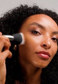 Femme aux cheveux bouclés appliquant un fond de teint liquide sur sa joue avec un pinceau de maquillage, teint de peau brun moyen.