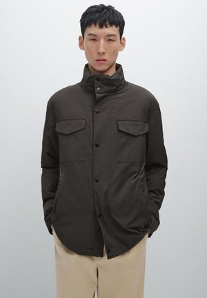 Donkerbruine jas met een hoge kraag, sluiting met knopen aan de voorkant, twee borstzakken en een gladde textuur, gecombineerd met beige broek.