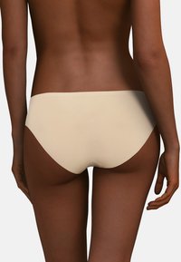 Beige sömlös hipsterunderkläder med slät textur, låg midja och rena linjer, placerad mot en mörk hudtonbakgrund.