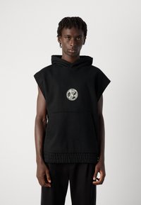 Young person with short dreadlocks wearing sleeveless black hoodie with circular white logo on the chest against plain background.