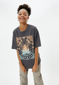 Maglietta grigia oversized con un design grafico multicolore che presenta elementi floreali e celestiali. Maniche corte e vestibilità comoda.