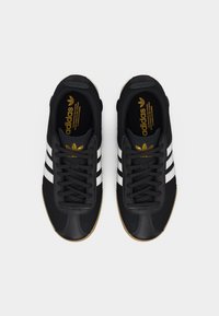 Ζευγάρι μαύρα αθλητικά παπούτσια Adidas με άσπρες ρίγες και καφέ σόλες, δεμένα με κορδόνια και φανερά από πάνω σε λευκό φόντο.
