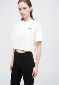 Femeie îmbrăcată cu un tricou scurt alb cu un logo mic și pantaloni negri cu talie înaltă, stând în fața unui fundal deschis simplu.