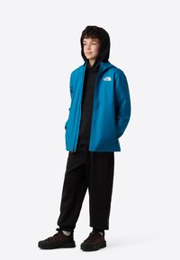 Jachetă albastră impermeabilă cu glugă, hanorac negru dedesubt, pantaloni negri și încălțăminte închisă la culoare, cu logo-ul The North Face pe piept.