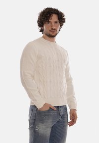 Uomo con capelli ricci che indossa un maglione a trecce crema e jeans blu strappati, in piedi con una mano in tasca contro uno sfondo neutro.