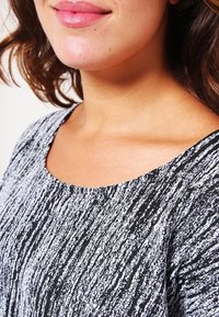 Femme portant un haut texturé noir et blanc, montrant sa partie inférieure du visage, son cou et ses épaules, avec des cheveux bruns ondulés.