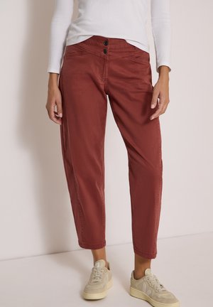 Femme portant un pantalon court taille haute rouge rouille avec fermeture à double bouton et des baskets beiges sur un fond uni.