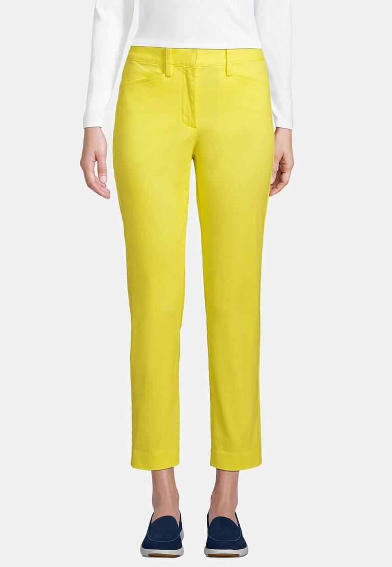 Femme portant un pantalon droit jaune vif, un haut blanc à manches longues et des chaussures bateau bleu marine, debout devant un fond uni.