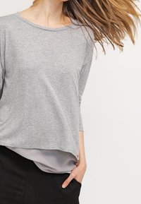 Nainen, jolla on pitkät hiukset, pukeutuneena kerrokselliseen harmaaseen toppiin ja mustiin housuihin, pitäen yhtä kättään taskussa yksinkertaista valkoista taustaa vasten.