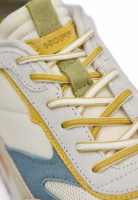 Zapatillas que combinan varios materiales: malla blanca, gamuza gris y detalles amarillos. Los cordones son amarillos y blancos con una etiqueta verde en la lengüeta.