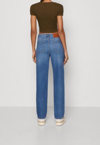 Blå raka jeans med medelblå tvätt, som har en brun läderpatch på baksidan, två fickor och en lös passform, kombinerat med en grön topp.