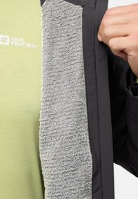 Sort jakke med en blød, grå fleeceforing; har en grøn skjorte nedenunder med et logo. Detaljen viser stoftekstur og lagdeling.