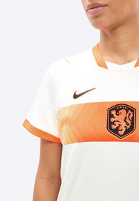 Άτομο που φοράει ποδοσφαιρική φανέλα λευκή και πορτοκαλί με λογότυπο Nike και το σύμβολο του λιονταριού της KNVB στο στήθος, με απλό ανοιχτό φόντο.