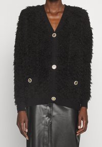 Schwarze strukturierte Strickjacke mit V-Ausschnitt, ausgestattet mit goldfarbigen Knopfverschlüssen und zwei Fronttaschen. Strickstoff mit flauschiger Optik.