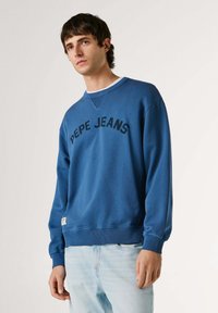 Jeune homme portant un sweat-shirt bleu Pepe Jeans et un jean bleu clair, debout devant un fond blanc uni.