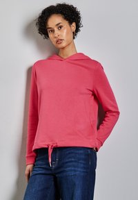 Sudadera rosa con cordón en la cintura, mangas largas y una textura suave. Llevada con jeans azules, mostrada contra un fondo neutro.