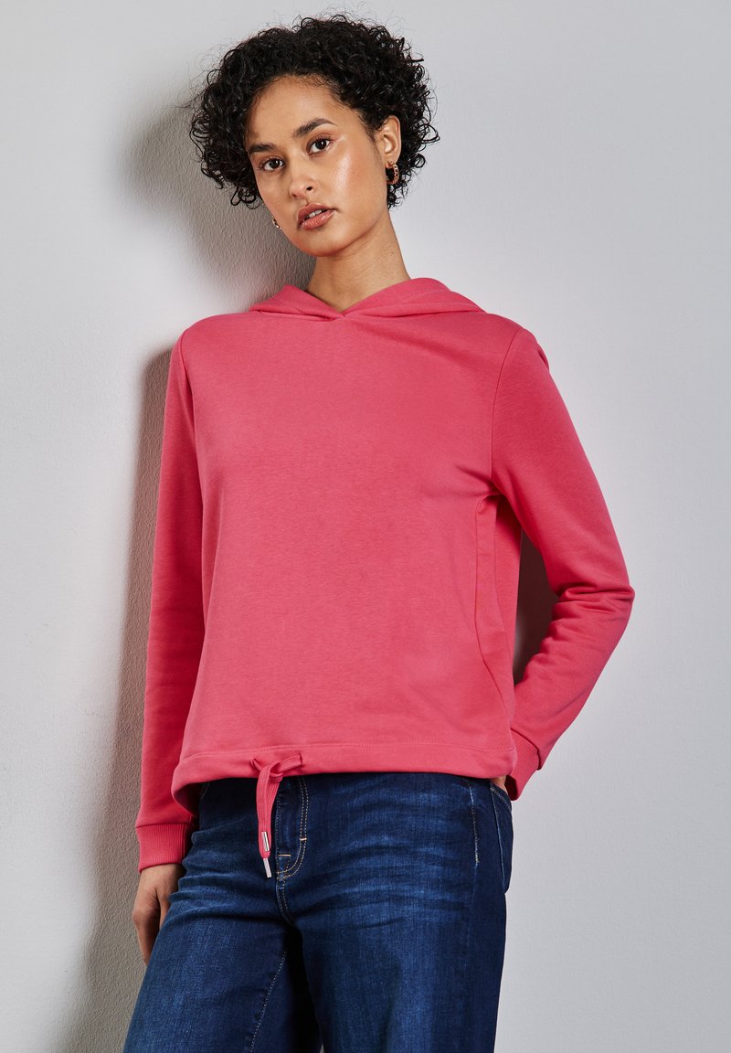 Sudadera rosa con cordón en la cintura, mangas largas y una textura suave. Llevada con jeans azules, mostrada contra un fondo neutro.