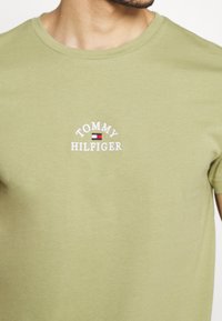 Camiseta de algodón color verde oliva, con un logo bordado en blanco de "TOMMY HILFIGER" y un detalle de bandera roja y azul en el pecho.