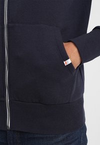 Tmavě modrá mikina na zip s kapsami vpředu. Má žebrované manžety a lem, s malou červenobílou štítkovou ozdobou na kapse.