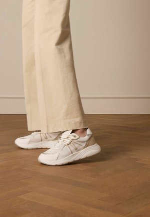 White sneakers with a textured finish, featuring gold hardware accents and a beige heel overlay, paired with light beige, wide-leg trousers.