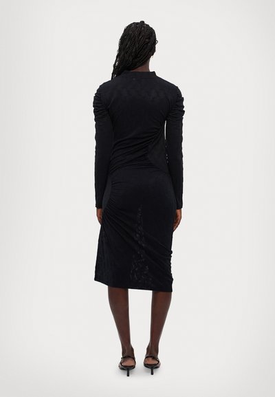 Zwarte jurk met lange mouwen van een textuur stof, met zijdelingse ruches en een slim-fit silhouet dat net onder de knie valt. Gladde afwerking met een lichte glans.