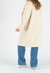 Crèmegekleurde, pluche, textuurgekleurde jas met een kraag, lange mouwen, twee zijzakken, gedragen over blauwe wijde jeans en witte sneakers.
