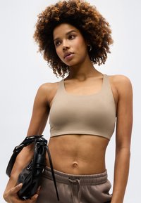 Νεαρή γυναίκα με σγουρά μαλλιά φορώντας μπεζ cropped τοπ και φαρδιά παντελόνια, κρατώντας μια μαύρη δερμάτινη τσάντα μπροστά σε μονόχρωμο φόντο.