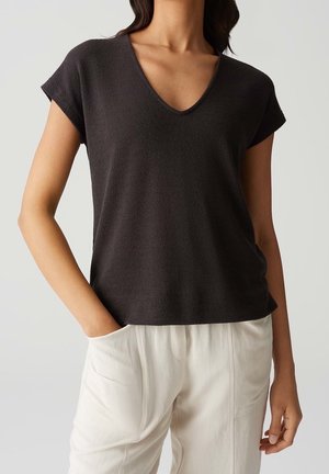 Femme portant un haut à manches courtes en V de couleur foncée et un pantalon clair, une main dans la poche, sur un fond uni.