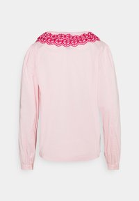 Blouse rose clair à manches longues avec épaules bouffantes et un large décolleté orné d'une bordure en dentelle florale rouge vif.