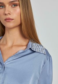 Camisa de satén azul claro con acentos de hombros adornados con tachuelas y un collar clásico, con una textura suave y diseño de botones en el frente.