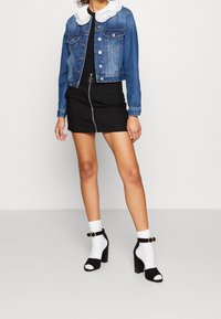 Giacca di denim con colletto in pizzo bianco, mini gonna nera con cerniera, tacchi con cinturino alla caviglia bianco e nero, calzini bianchi con risvolto, sfondo semplice.