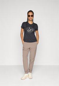 Kvinne med flettet hår ikledd solbriller, svart grafisk t-skjorte, beige joggebukser og hvite sneakers som står med hendene i lomma.