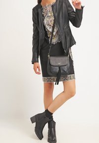 Femme portant une veste en cuir noire et des bottines, une robe noire à motifs, et un sac bandoulière noir avec des pompons.