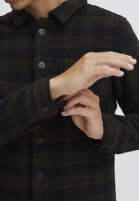 Black and brown plaid shirt jacket, made of a textured fabric with a button front and cuffs, showcasing a close-up of hands adjusting the sleeve.