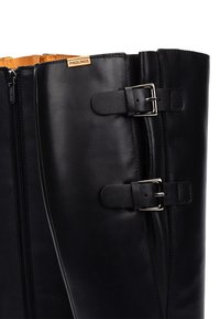Bota de cuero negro hasta la rodilla con acentos de hardware plateado, dos correas con hebillas y un zipper en el lateral. Textura suave con un acabado ligeramente brillante.