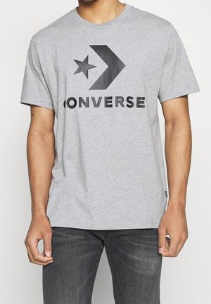 Mężczyzna ubrany w szarą koszulkę z krótkim rękawem z dużym czarnym logo Converse oraz ciemnoszare dżinsy, stojący na tle jednolitej ściany.